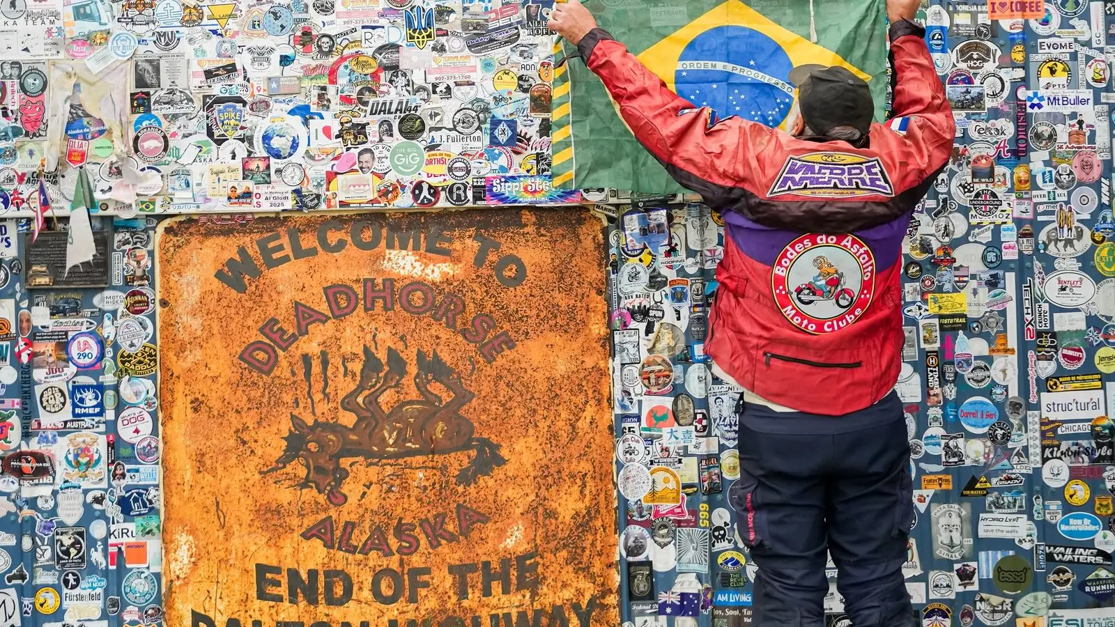 Piloto ergue a bandeira do Brasil diante da placa “Welcome to Deadhorse — End of the Dalton Highway”, em Prudhoe Bay